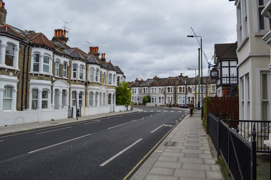 A quiet residential street with rows of Victorian-style terraced houses featuring white facades, bay windows, and red tiled roofs situated on the left side, while on the right side there are modern white buildings with black trim and balconies. The street is paved with asphalt and has clearly marked parking spaces and a sidewalk made of concrete slabs, with a few pedestrians walking along. Overhead, there are streetlights and a large crane visible in the background, indicating ongoing building work. The sky is overcast with grey clouds. This setting is typical of an urban neighbourhood in Kilburn, NW6, where house removals and furniture transport services by Man with Van Kilburn might be carried out as part of home relocation or moving logistics.