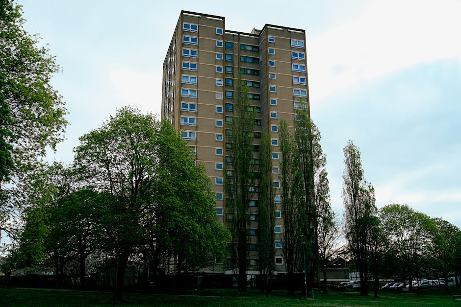 A tall, multi-storey residential building with a flat roof and numerous windows, surrounded by tall, leafy trees in a green outdoor area. The building’s exterior is constructed from grey concrete, with blue window frames. The environment appears to be a residential neighbourhood, with a paved area at the building’s base and parked cars visible in the background. The scene is lit by natural daylight, and the image captures the building’s height from a low-angle perspective, emphasizing the urban setting. This image relates to house removals and furniture transport, illustrating the type of property involved in home relocation services, with the surrounding environment providing context for professional storage and moving logistics.