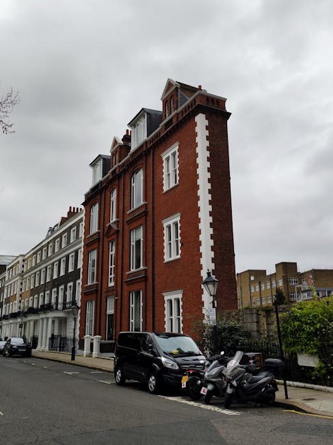 A quiet residential street with rows of Victorian-style terraced houses featuring white facades, bay windows, and red tiled roofs situated on the left side, while on the right side there are modern white buildings with black trim and balconies. The street is paved with asphalt and has clearly marked parking spaces and a sidewalk made of concrete slabs, with a few pedestrians walking along. Overhead, there are streetlights and a large crane visible in the background, indicating ongoing building work. The sky is overcast with grey clouds. This setting is typical of an urban neighbourhood in Kilburn, NW6, where house removals and furniture transport services by Man with Van Kilburn might be carried out as part of home relocation or moving logistics.