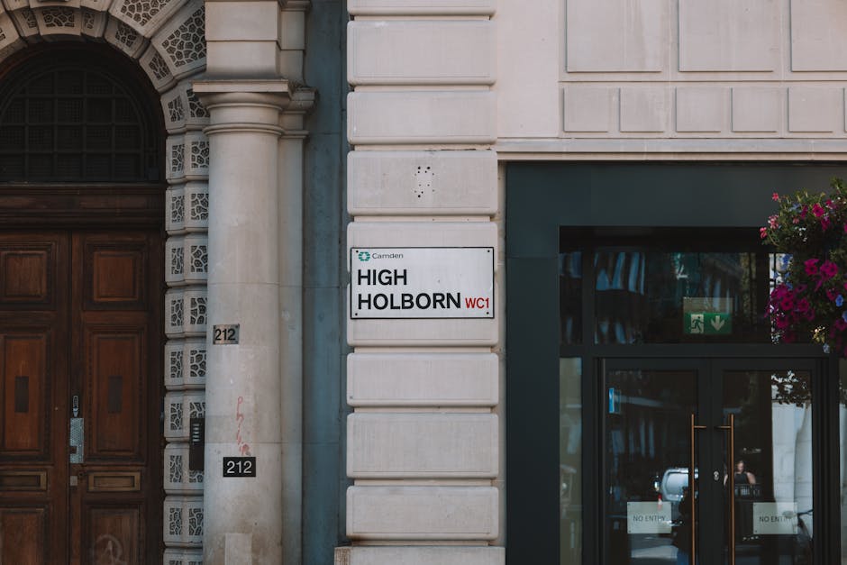 Close-up view of a building façade in an urban setting showing a white street sign with black and red text that reads 'High Holborn WC1' attached to the corner of a beige stone wall. To the left of the sign, there is a large, dark wooden door with decorative carvings, and a small black intercom panel mounted nearby. To the right, part of a glass entrance door with a metal handle is visible, featuring a green emergency exit sign with a white arrow and figure inside the glass. Above the door, there are hanging flower baskets with vibrant pink and purple flowers. The scene appears to be part of a home or commercial property, and the overall environment suggests a location suitable for professional house removals and furniture transport, with attention to details such as packaging materials or moving equipment not visible in this shot. The image captures the environment's natural lighting, emphasizing the contrast between the traditional door and modern signage, illustrating the typical setting for house relocation processes facilitated by [COMPANY_NAME].