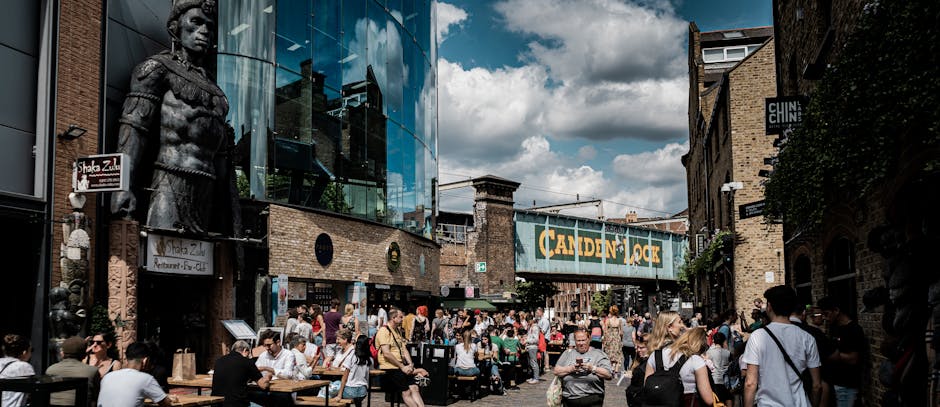 A busy street scene outside a restaurant and retail establishments in Kilburn, with numerous pedestrians walking and sitting at outdoor tables, some holding food and beverages. In the background, a modern glass-fronted building displays large decorative sculptures, including a statue of a historical figure mounted on the exterior wall. A prominent overhead sign reads 'CANDEN ROCK,' indicating local signage. The scene suggests active urban surroundings typical of a lively area for house relocations and moving services, with pedestrians and outdoor furniture near the pavement, and a partly cloudy sky overhead. The overall environment is bright and bustling, reflecting the social and commercial atmosphere relevant to home relocation and furniture transport activities in Kilburn, supported by local removals company Man with Van Kilburn.