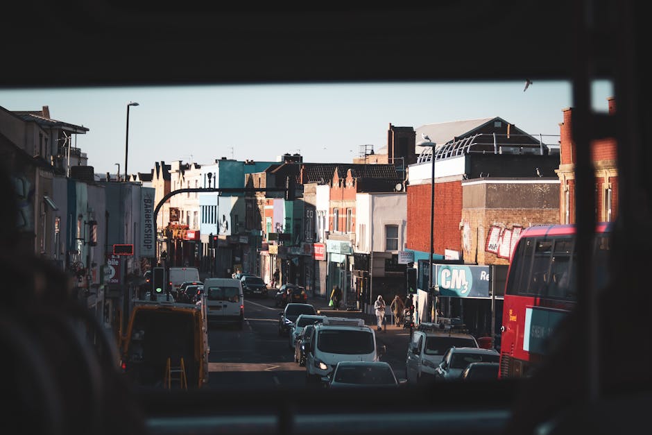 View of a busy urban street captured from inside a moving vehicle, with the image framed by the vehicle's window. The street is lined with various small shops and commercial buildings with different coloured facades and signage, including a barber shop and other retail stores. Several cars, vans, and a red double-decker bus are visible on the road, with some vehicles waiting at a traffic light. Pedestrians are crossing the street and walking along the sidewalks. The area appears to be in the midst of a home relocation effort, with the scene illustrating the typical moving logistics involving vehicle transport and street-based loading and unloading processes, which [COMPANY_NAME] offers as part of their removals service coverage in Kilburn. Bright daylight illuminates the scene, highlighting the everyday activity associated with house moves and furniture transport within an urban environment.