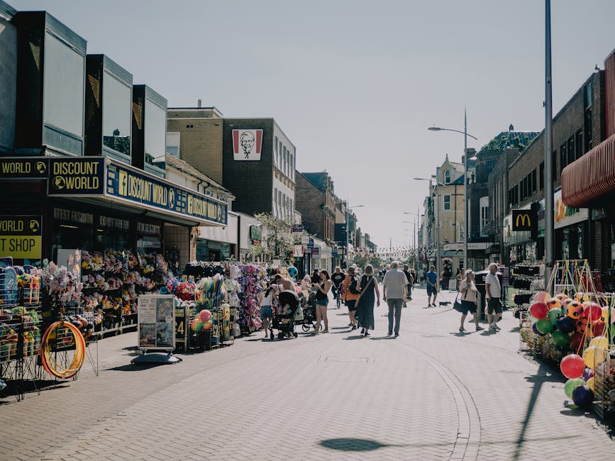 A busy pedestrians' shopping street in Kilburn during daytime, with storefronts on both sides displaying various items such as flowers, balloons, and toys. People are walking, pushing strollers, and browsing shops, some engaged in conversations. The street is lined with commercial buildings, including a discount store with signs advertising fashion and equipment. The scene features outdoor displays with colorful merchandise placed on shelves and racks outside the shops, and a vehicle, possibly a van used for furniture transport or packing during house removals, partially visible to the right side. The environment is illuminated with natural daylight, casting shadows on the paved street, reflecting typical elements of local home relocation logistics and furniture moving activities associated with professional removals services like those offered by Man with Van Kilburn.