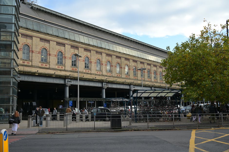 The image shows the exterior of Kilburn Park Station with a multi-storey brick and stone building featuring arched windows. In front of the station, a group of people are gathered on the pavement, some standing and others using a plastic-covered bike rack filled with bicycles. A disabled parking sign is visible near the curb, with several parked cars lining the street. To the left, part of a modern glass building can be seen, contrasting with the historic station structure. The scene includes a covered bike storage area adjacent to the station, and trees with green leaves are present on the right side of the image. The overall environment suggests an active street scene during daytime, with clear weather, suitable for home relocation and furniture transport activities conducted by services such as Man with Van Kilburn. The image captures the logistics involved in loading or unloading items during a house removal process, with attention to the environment and surroundings relevant to moving services in Kilburn.