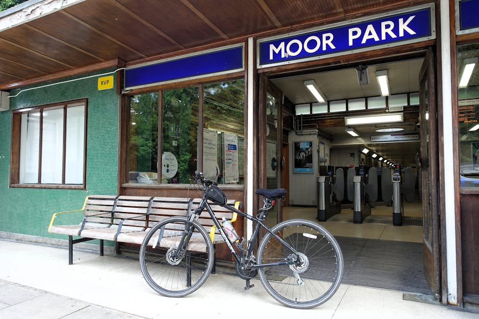 The image shows the exterior of Kilburn Park Station with a multi-storey brick and stone building featuring arched windows. In front of the station, a group of people are gathered on the pavement, some standing and others using a plastic-covered bike rack filled with bicycles. A disabled parking sign is visible near the curb, with several parked cars lining the street. To the left, part of a modern glass building can be seen, contrasting with the historic station structure. The scene includes a covered bike storage area adjacent to the station, and trees with green leaves are present on the right side of the image. The overall environment suggests an active street scene during daytime, with clear weather, suitable for home relocation and furniture transport activities conducted by services such as Man with Van Kilburn. The image captures the logistics involved in loading or unloading items during a house removal process, with attention to the environment and surroundings relevant to moving services in Kilburn.