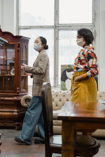 Two women wearing face masks are inside a room with large windows, engaged in a home relocation or packing process. One woman, dressed in a checkered blazer and jeans, is standing near a dark wooden display cabinet, possibly organizing or preparing items for removal. The other woman, wearing a colorful plaid shirt and mustard yellow trousers, is standing nearby, observing or assisting. The room contains a cream tufted sofa, a wooden table, and a painting of a horse leaning against the wall. Natural light filters through the expansive windows, illuminating the area. The scene suggests a furniture transport or packing and moving activity, with the environment showing typical household furniture and a professional approach to house removals, consistent with services offered by Man with Van Kilburn.
