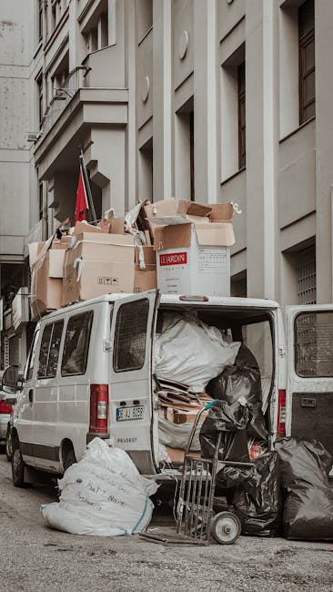 A white panel van parked on a city street with its rear doors open, revealing various packing materials, including large black and white plastic bags, cardboard boxes, and packing paper inside. Several oversized cardboard boxes, some partially collapsed, are stacked on top of the van, secured with packing tape. A small hand truck or dolly is positioned on the pavement next to the van, loaded with additional boxes and bags. The van is situated in front of a multi-storey building with beige or light grey exterior walls, windows, and architectural details. The scene appears to depict the loading or unloading process involved in a house removal or relocation, consistent with professional moving services such as those offered by Man with Van Kilburn.