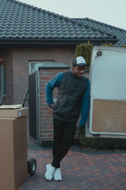 A man wearing a black cap, white T-shirt, and jeans is placing three cardboard boxes into the side door of a white panel van parked on a residential street. The van, used for home relocation or furnit