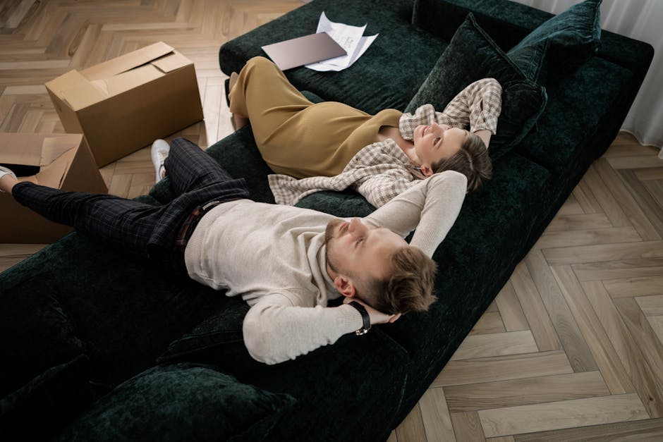 A young woman and a young man are lying on a dark green velvet sofa inside a living room, appearing relaxed and smiling at each other. The woman is wearing a plaid shirt and beige skirt, with her head resting on the sofa's armrest and her left arm extended behind her head. The man is dressed in a cream sweater and dark checkered trousers, lying on his back with his head on the sofa, facing the woman. There are several cardboard boxes placed on the wooden floor nearby, some open with packing materials inside, indicating a home relocation process. A few sheets of paper are placed on the sofa beside the woman. The room has natural lighting, and the scene captures a moment during packing and moving, with furniture and packaging materials set around, suggesting an ongoing move managed by professionals like Man with Van Kilburn.