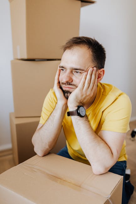 A man with short dark hair and a beard, wearing a yellow T-shirt and a black wristwatch, is seated at a table inside a room filled with cardboard moving boxes. He is resting his head in his hands, looking stressed or overwhelmed. Behind him, several stacked cardboard boxes of various sizes are visible, some sealed with packing tape. The scene suggests a home relocation or packing process associated with house removals. The environment appears well-lit with natural or artificial light, and there is no visible furniture apart from the boxes. This image aligns with the context of furniture transport, packing, and moving logistics, relevant to the services offered by Man with Van Kilburn in house removals.